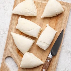 Six pieces of dough are placed on a wooden cutting board next to a knife.