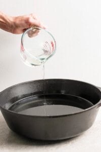 Hand pouring liquid from a measuring cup into a black cast iron pan.