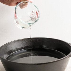 Hand pouring liquid from a measuring cup into a black cast iron pan.