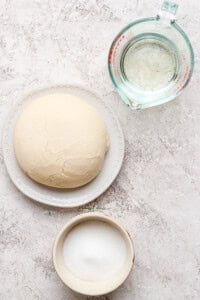 Dough ball on a plate, a measuring cup with water, and a bowl of sugar on a textured surface.