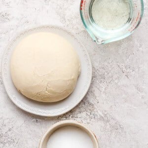 Dough ball on a plate, a measuring cup with water, and a bowl of sugar on a textured surface.