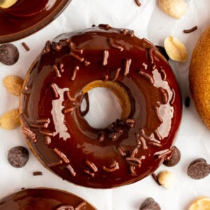 A chocolate-glazed donut with chocolate sprinkles, surrounded by chocolate chips and peanuts on a white surface.