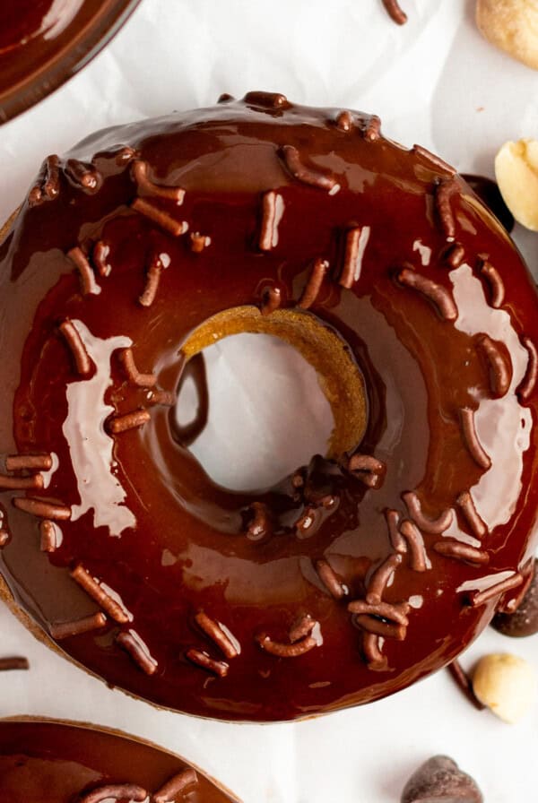 A chocolate-glazed donut with chocolate sprinkles, surrounded by chocolate chips and peanuts on a white surface.