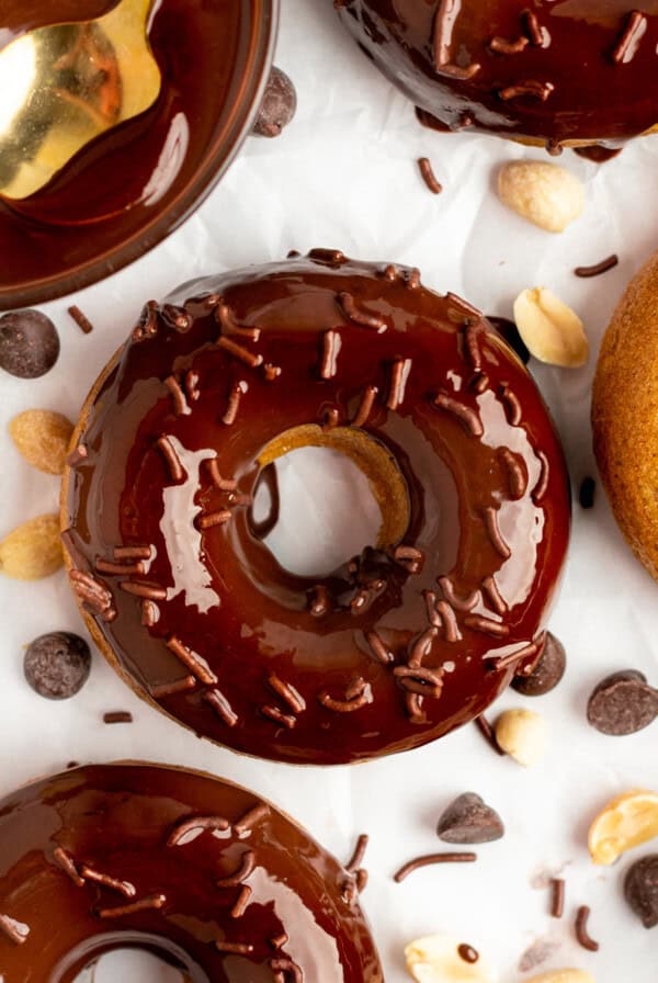 Close-up of chocolate-glazed donuts topped with chocolate sprinkles, surrounded by peanuts and chocolate chips on a white background.