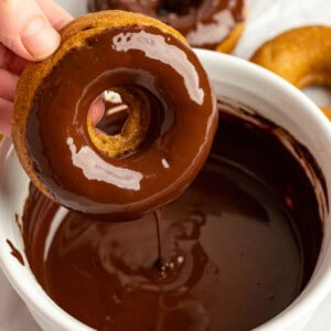 A hand dips a donut into a bowl of chocolate glaze, with more donuts on a white surface in the background.