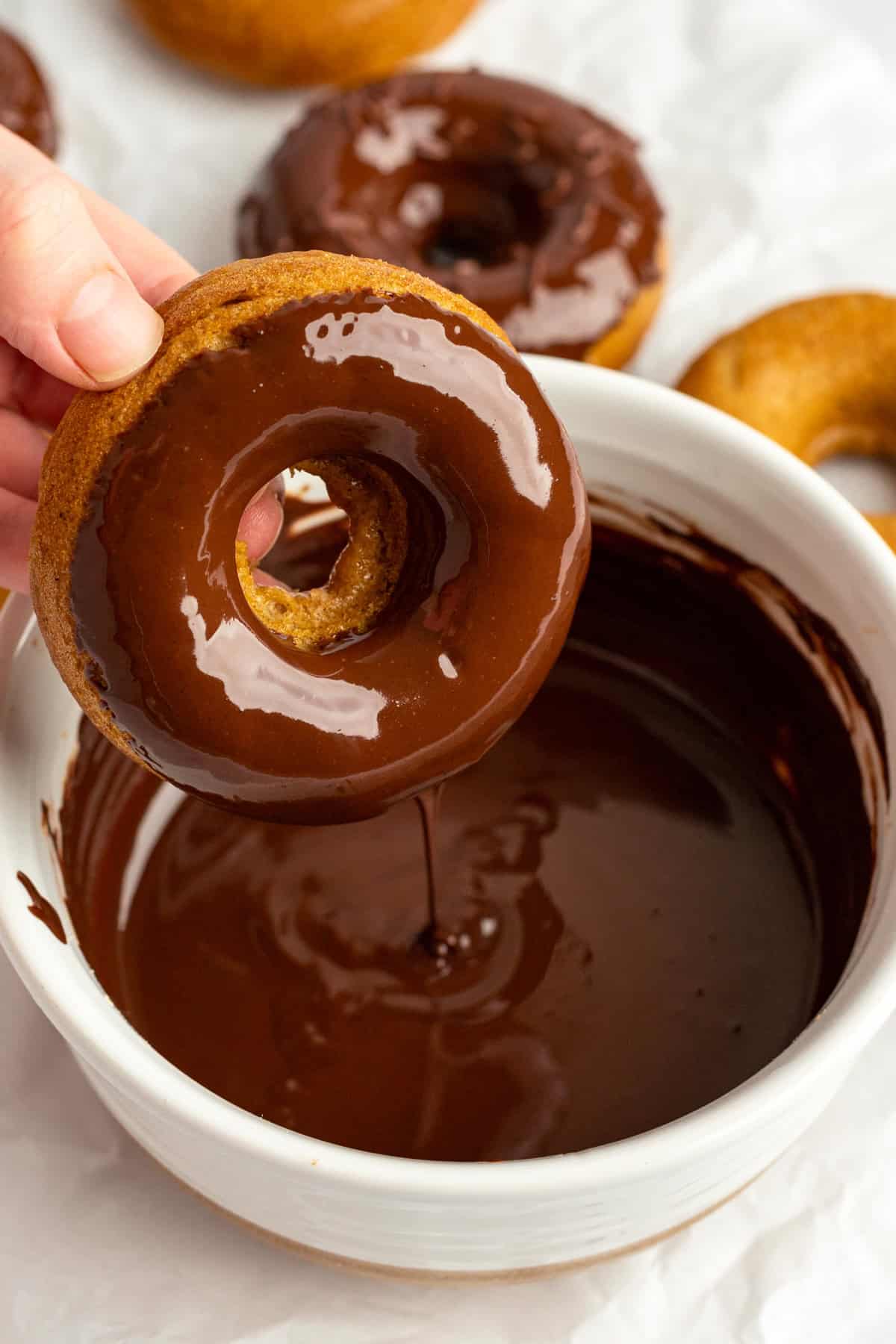 A hand dips a donut into a bowl of chocolate glaze, with more donuts on a white surface in the background.