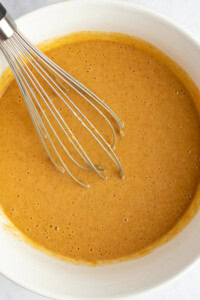 A metal whisk in a white bowl containing smooth, light brown cake batter.