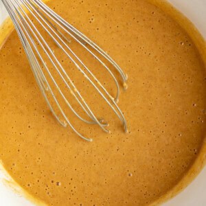 A metal whisk in a white bowl containing smooth, light brown cake batter.