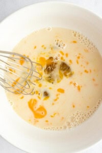 A whisk mixing eggs, milk, and oil in a white bowl.