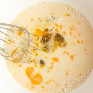 A whisk mixing eggs, milk, and oil in a white bowl.