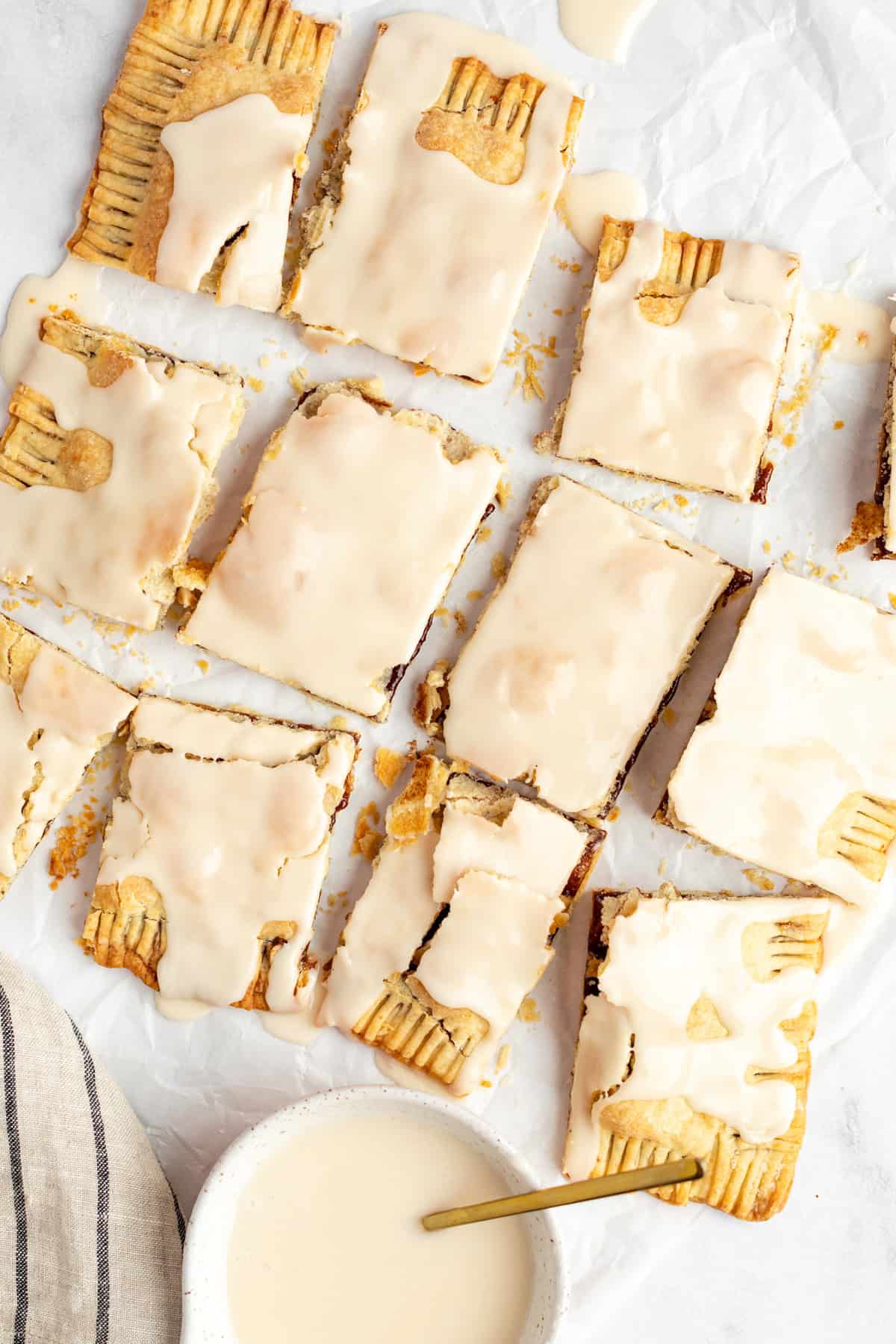 Nine iced pastry rectangles with a crumbly texture, arranged on parchment paper. A bowl of icing with a spoon is at the bottom left.