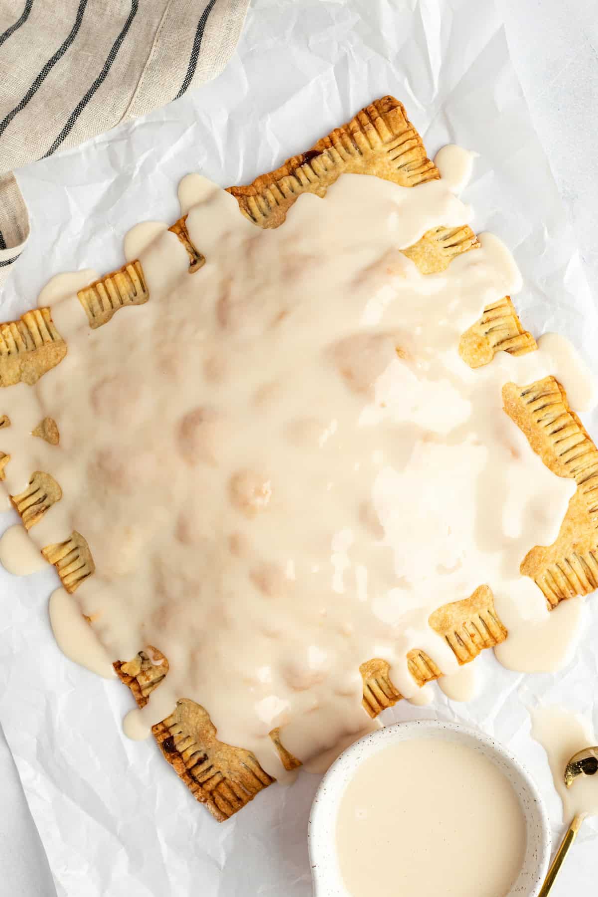 A large rectangular pastry with ridged edges is covered in white icing, placed on crinkled parchment paper next to a small bowl of extra icing.