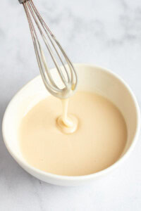A whisk with creamy batter drips into a white bowl on a light surface.