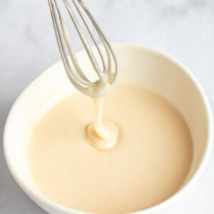 A whisk with creamy batter drips into a white bowl on a light surface.