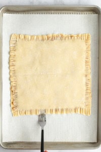 A fork is pressing down the edges of a rectangular pastry on parchment paper in a baking tray.