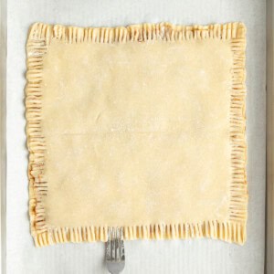 A fork is pressing down the edges of a rectangular pastry on parchment paper in a baking tray.