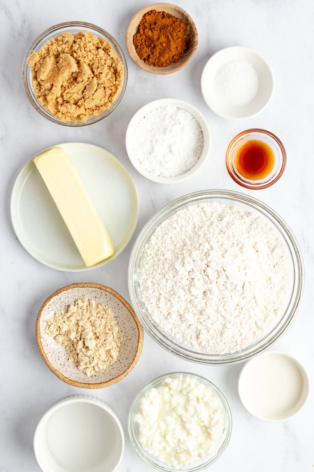 Assorted baking ingredients on a white surface: flour, brown sugar, butter, cocoa powder, baking powder, vanilla extract, sour cream, and milk in bowls and plates.