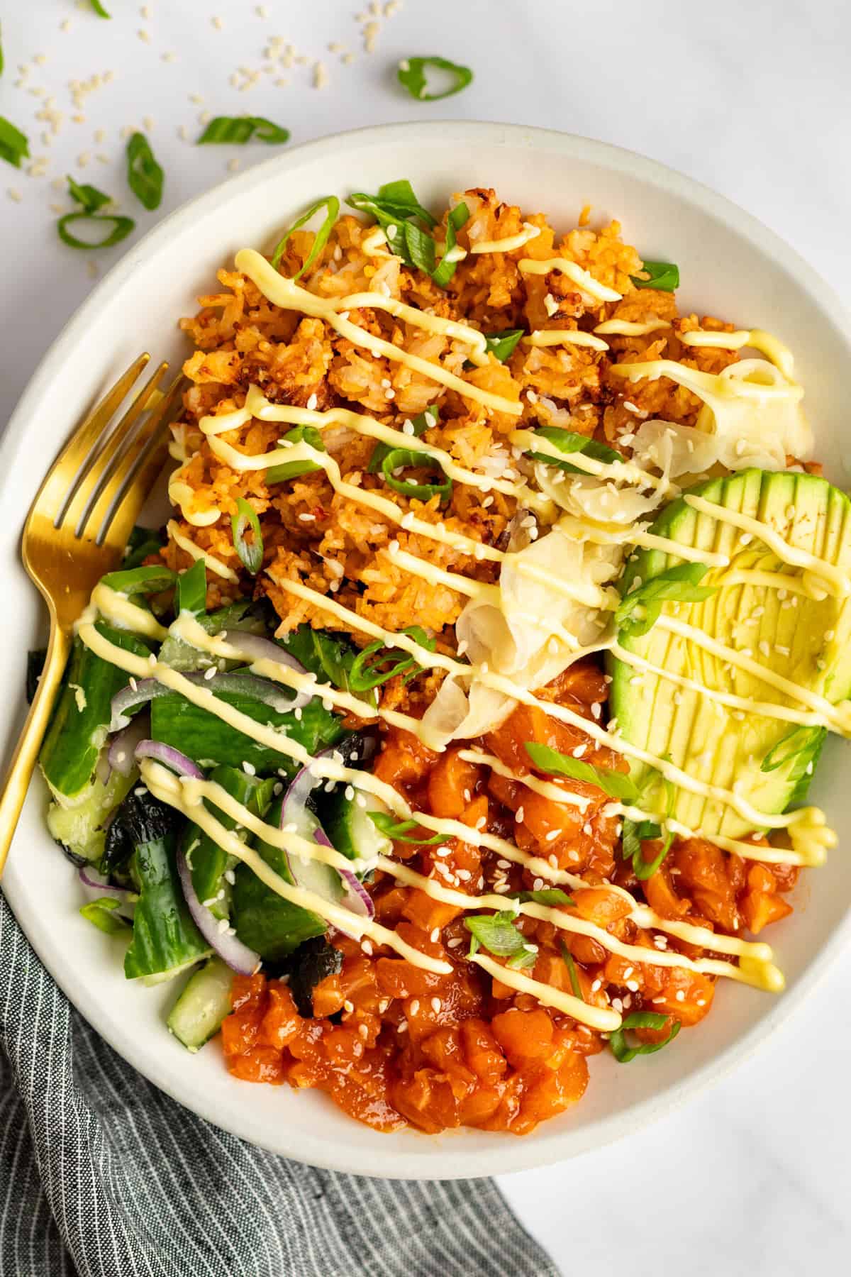 A bowl of rice topped with avocado slices, marinated raw fish, sliced vegetables, shredded cabbage, and drizzled with a creamy sauce. A gold fork rests on the side.