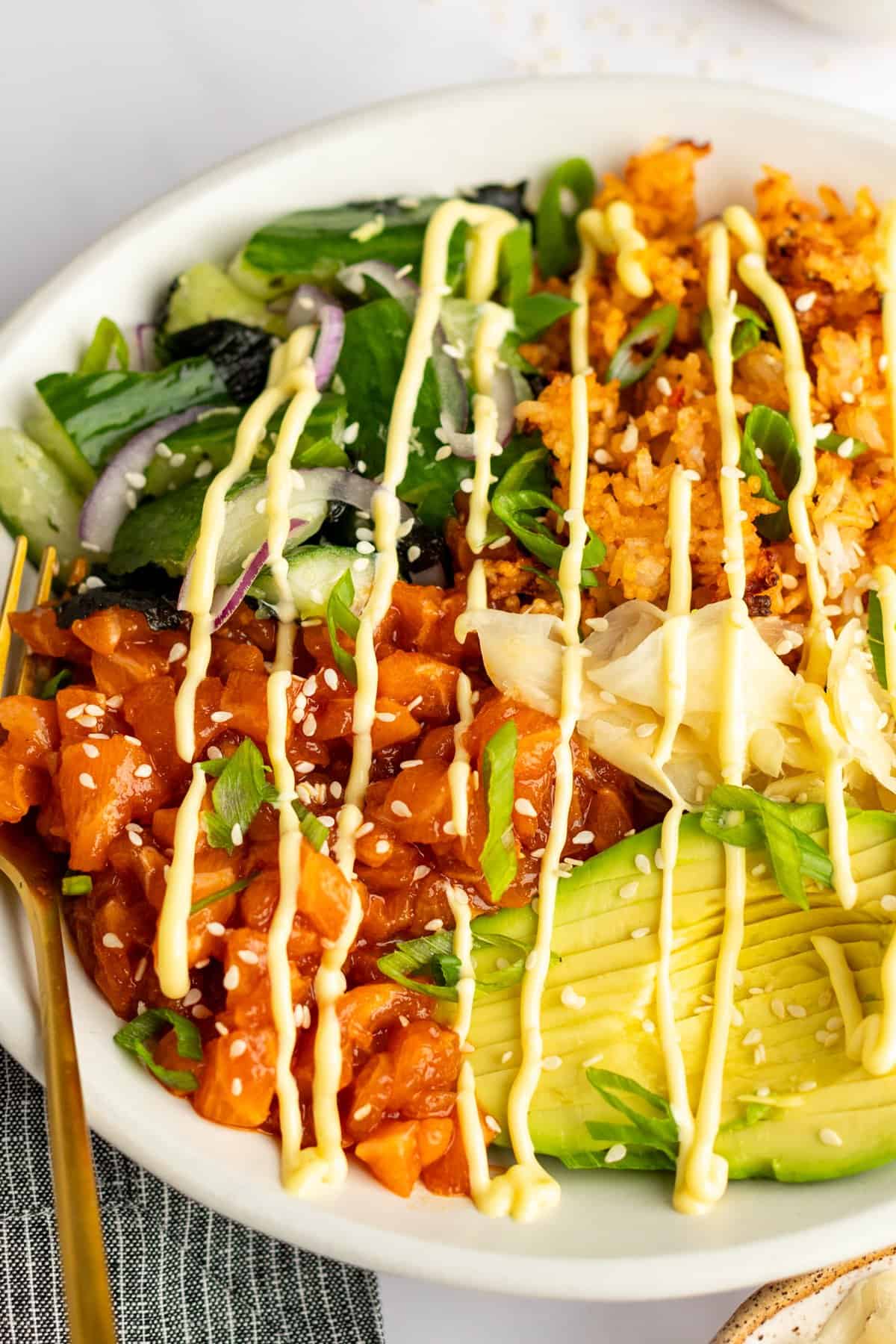 A bowl with diced salmon, avocado slices, greens, onions, rice, and mayo drizzle, garnished with sesame seeds and scallions.