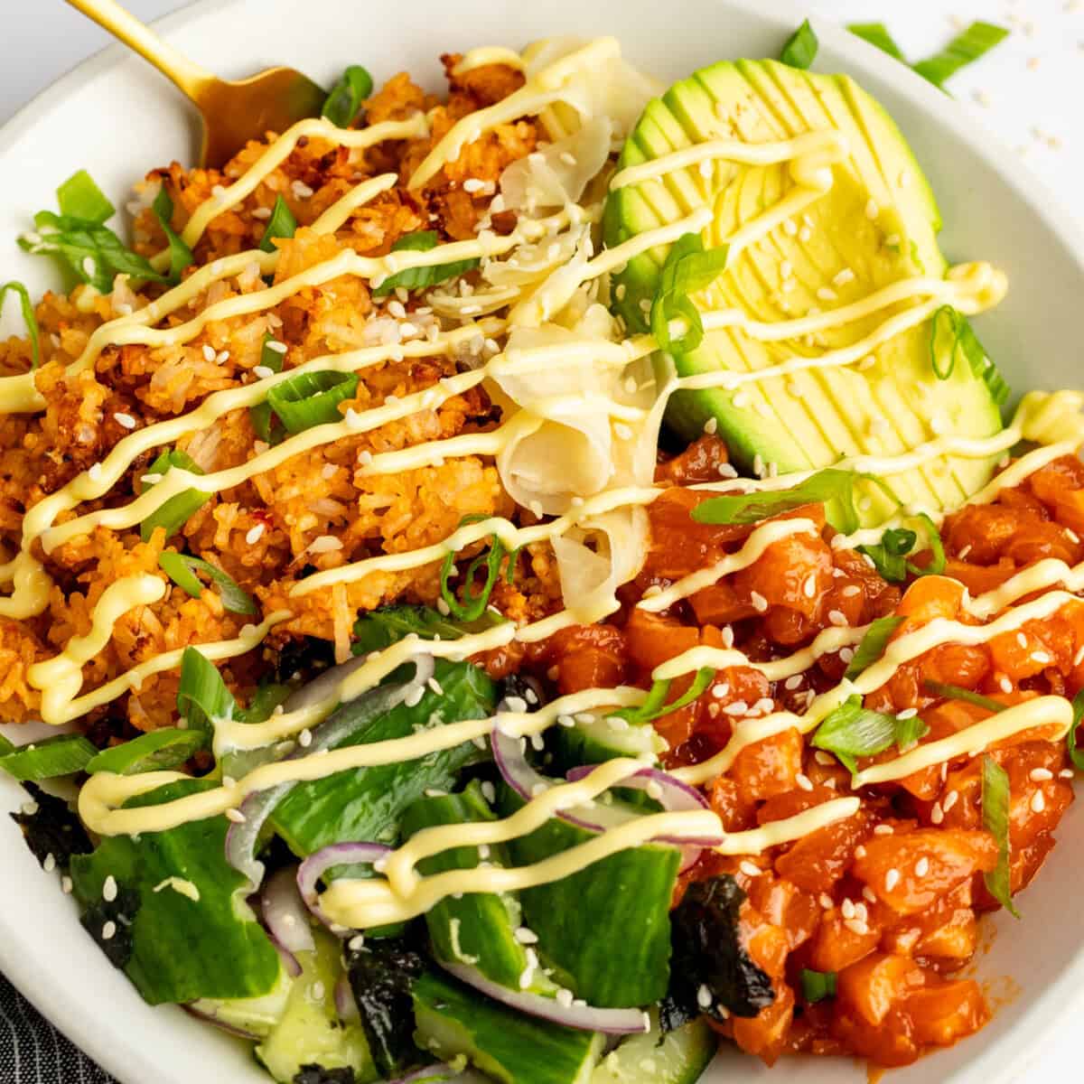 A bowl with rice, avocado slices, diced fish, greens, and seaweed, topped with mayonnaise drizzle and sprinkled with sesame seeds and green onions.