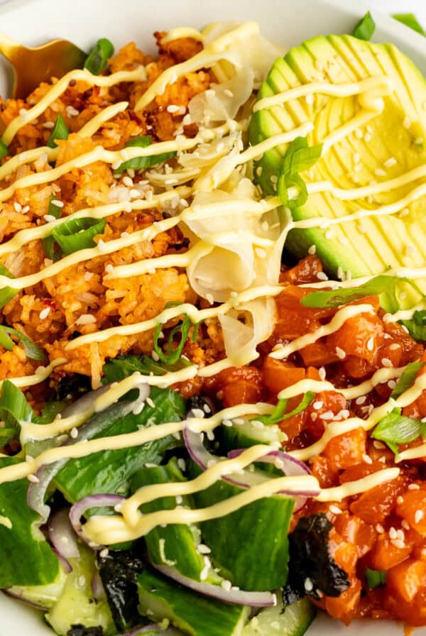 A bowl with rice, avocado slices, diced fish, greens, and seaweed, topped with mayonnaise drizzle and sprinkled with sesame seeds and green onions.