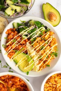 A bowl of rice topped with avocado, kimchi, salad greens, drizzled with mayonnaise, next to half an avocado and additional side dishes.
