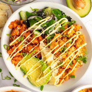 A bowl of rice topped with avocado, kimchi, salad greens, drizzled with mayonnaise, next to half an avocado and additional side dishes.