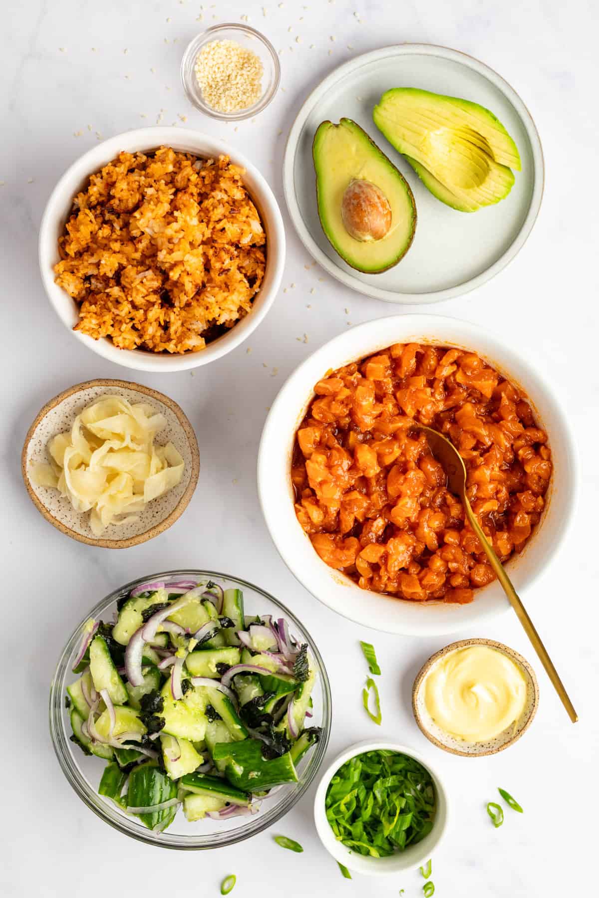 Ingredients for poke bowl on a white surface: raw salmon, sliced avocado, rice, cucumber salad, pickled ginger, sliced onions, mayonnaise, sesame seeds, and chopped green onions.