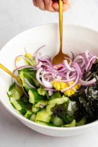A hand mixes a salad in a white bowl with a gold spoon. The salad contains sliced cucumbers, red onion, seaweed, and a dollop of mustard.