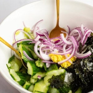 A hand mixes a salad in a white bowl with a gold spoon. The salad contains sliced cucumbers, red onion, seaweed, and a dollop of mustard.