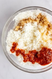 A bowl containing white rice, crumbled cheese, soy sauce, and red chili sauce on a light surface.