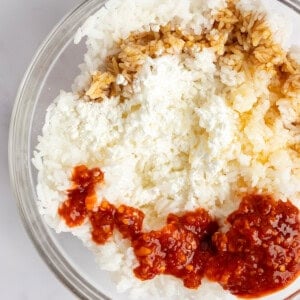 A bowl containing white rice, crumbled cheese, soy sauce, and red chili sauce on a light surface.