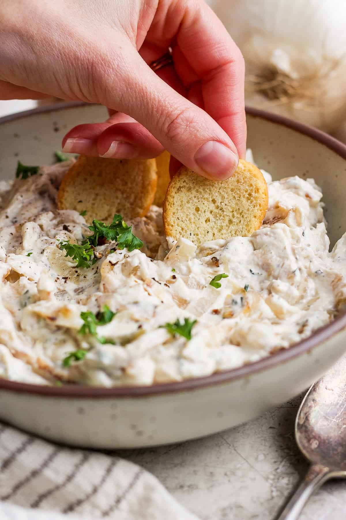 A hand dipping a small round cracker into a bowl of creamy dip garnished with parsley.