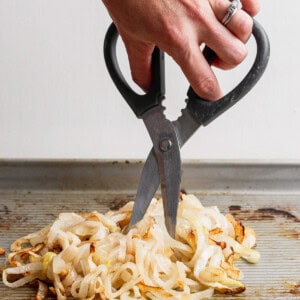 A hand using kitchen scissors to cut cooked onions on a baking sheet.