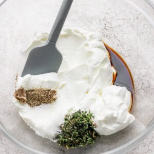 A mixing bowl with yogurt, soy sauce, herbs, and spices, stirred with a gray spatula.