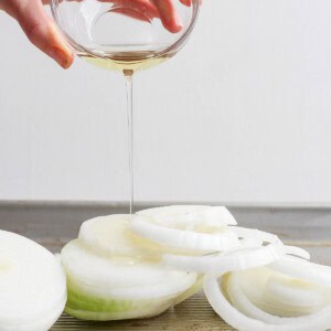 A hand pours olive oil from a glass bowl onto sliced onions on a baking sheet.