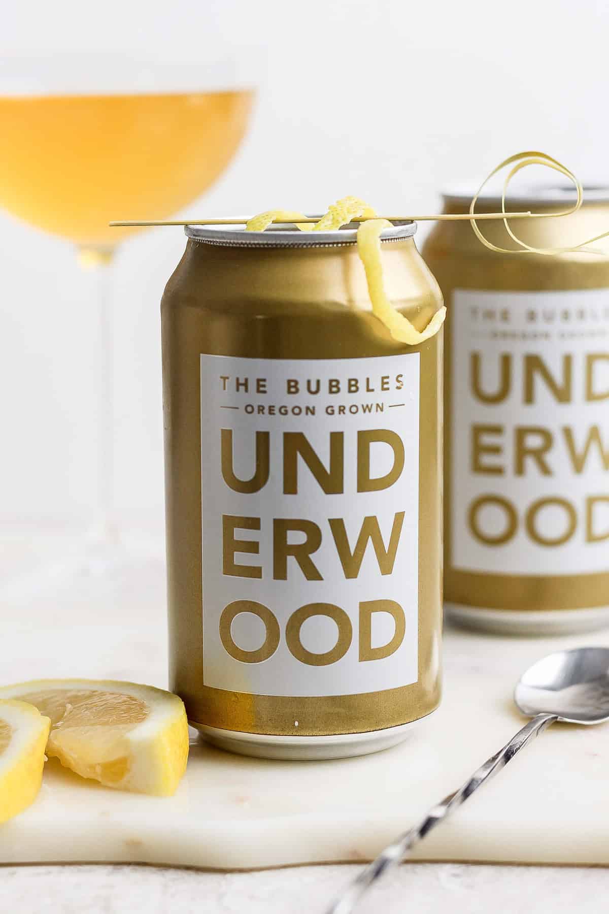 Two cans of Underwood "The Bubbles" canned champagne cocktail with lemon garnish grace the table, accompanied by a spoon and lemon slices. A wine glass adds elegance in the background.