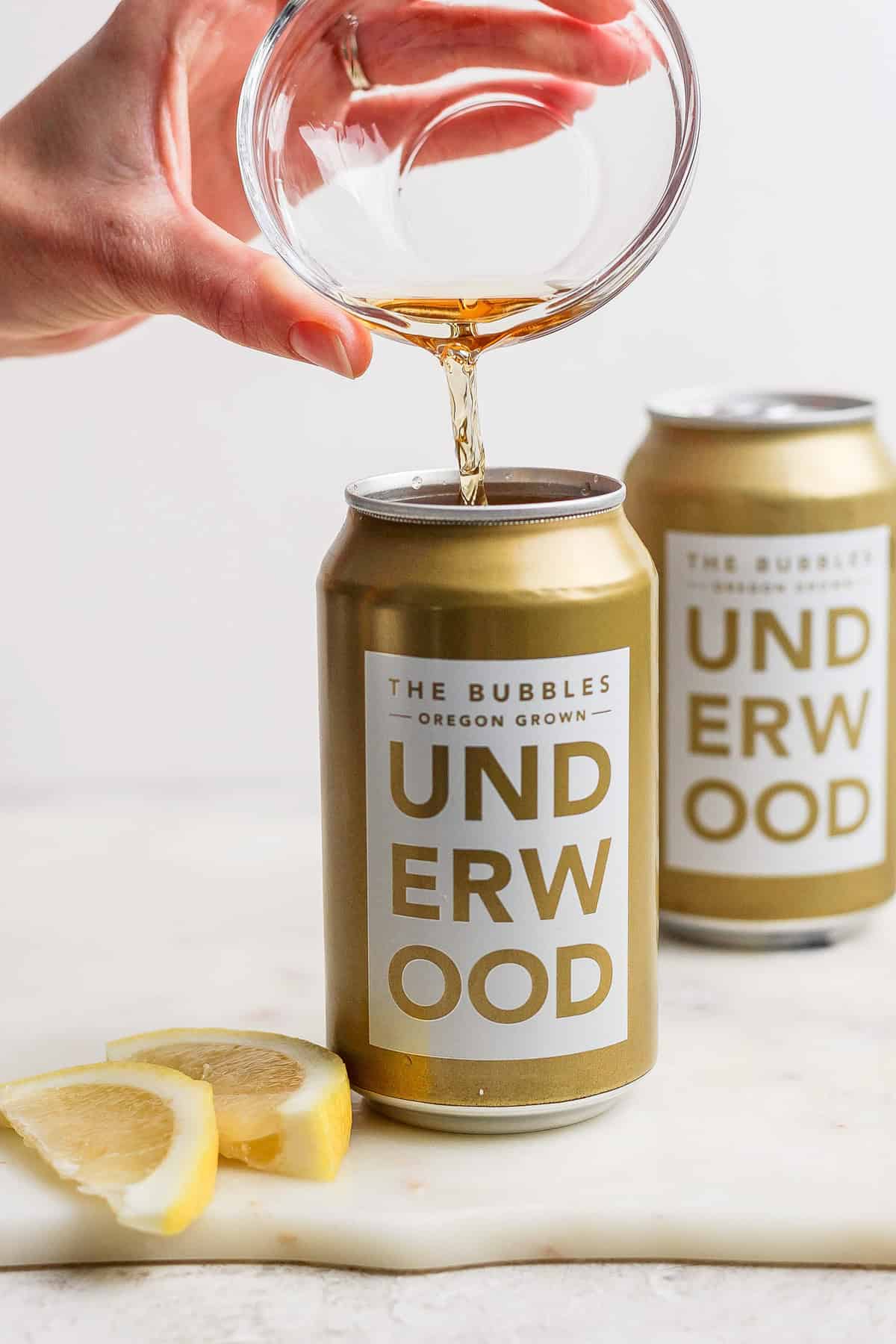 A hand elegantly pours liquid from a glass into a can labeled "Underwood: The Bubbles Oregon Grown," creating the perfect canned champagne cocktail. Two lemon slices and another can are subtly visible in the background, adding a touch of zest to this sparkling scene.