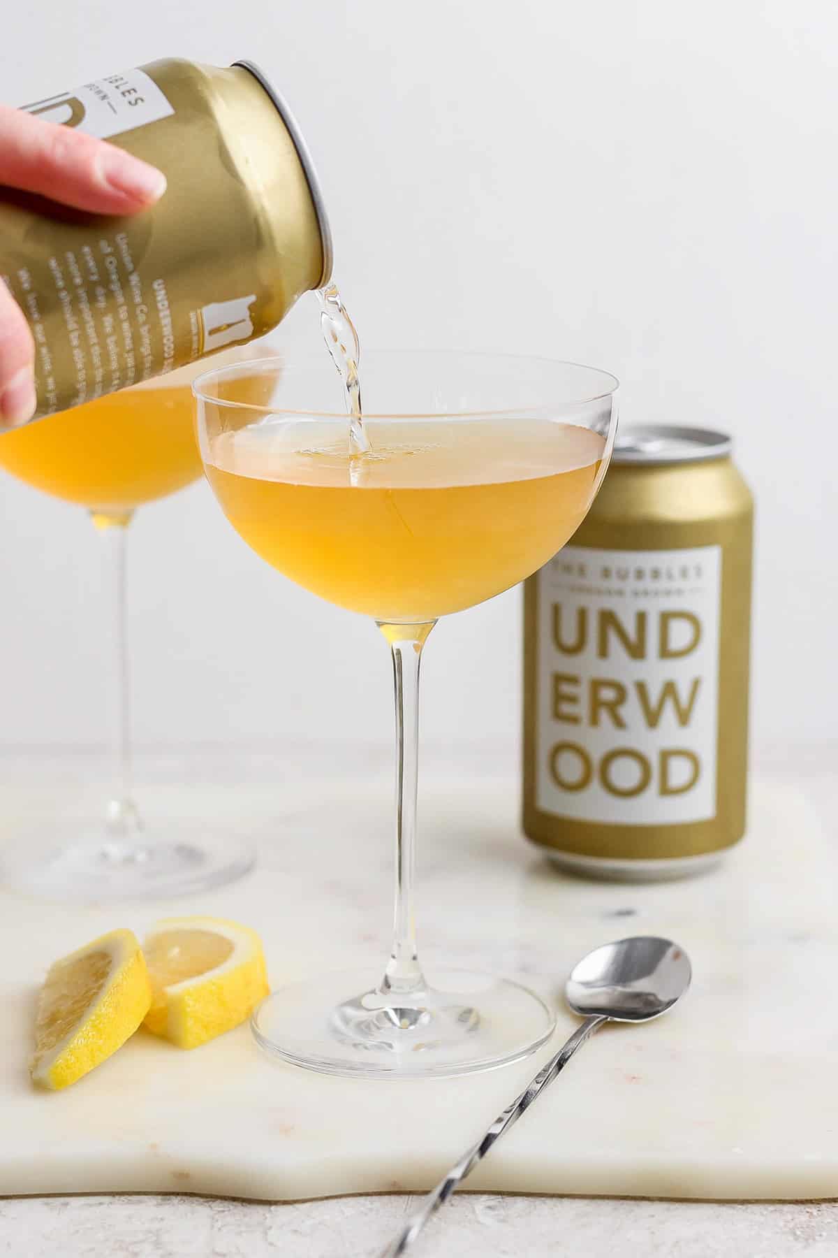 A can labeled "Underwood" is pouring a golden liquid, reminiscent of a canned champagne cocktail, into a coupe glass. Another filled glass, the can itself, lemon wedges, and a spoon are arranged on the marble surface.