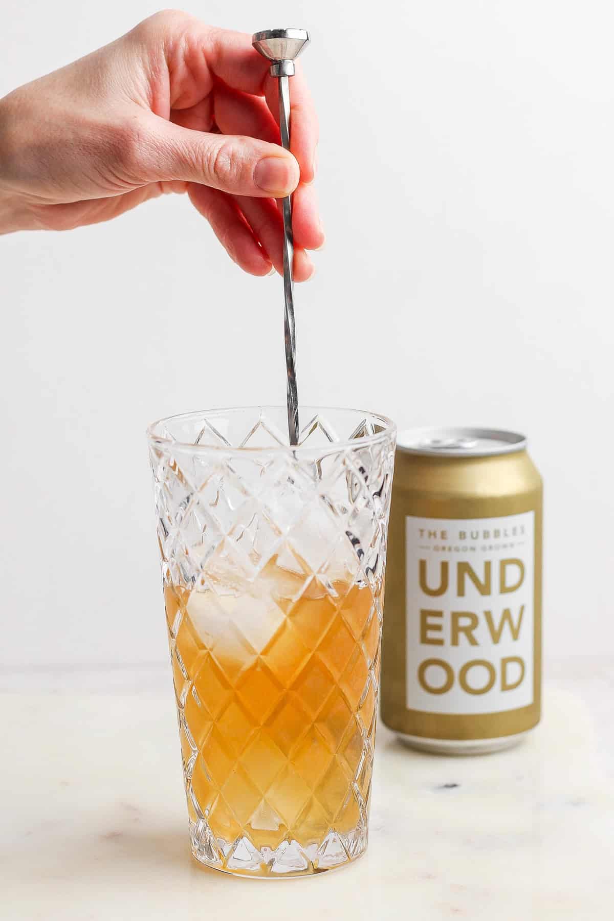 A hand stirs a canned champagne cocktail with ice in a diamond-patterned glass, while an unopened can of Underwood sparkling wine sits elegantly in the background.