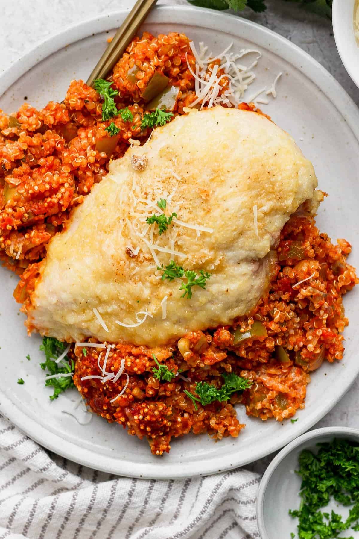 Grilled chicken breast on a bed of tomato quinoa, garnished with grated cheese and parsley on a white plate.