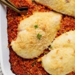 Baked chicken breasts with cheese on a bed of tomato sauce, garnished with parsley and grated cheese, in a white dish.