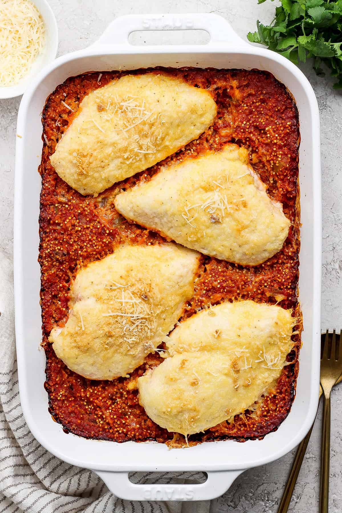 Baked chicken breasts topped with cheese in a casserole dish, resting on a red sauce base, with a fork and cloth napkin beside it.