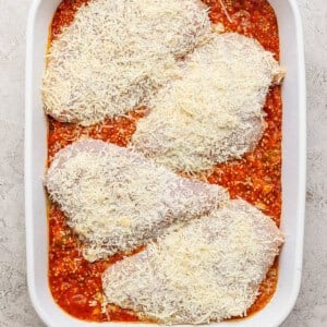 Four uncooked chicken breasts covered in grated cheese are placed on top of a layer of tomato sauce in a white baking dish.