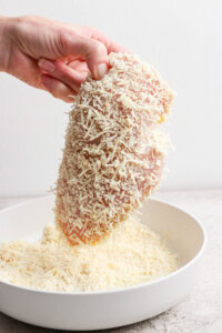 A hand holding a raw chicken breast coated with grated cheese over a white bowl.