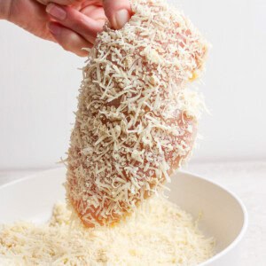 A hand holding a raw chicken breast coated with grated cheese over a white bowl.