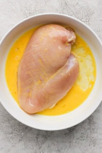 Raw chicken breast in a white bowl with beaten eggs on a gray textured surface.