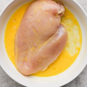 Raw chicken breast in a white bowl with beaten eggs on a gray textured surface.