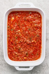 Close-up of a white baking dish filled with red tomato sauce mixed with visible herbs and spices.