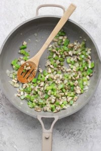 A pan with diced green bell pepper, onion, and mushroom being sautéed with a wooden spoon.